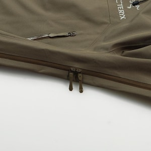 May include: Close-up of a khaki-colored jacket with a zipper closure and a side pocket. The jacket features the brand name "Arc'teryx" and the "Gore-Tex" logo. The jacket is made of a durable, waterproof material.
