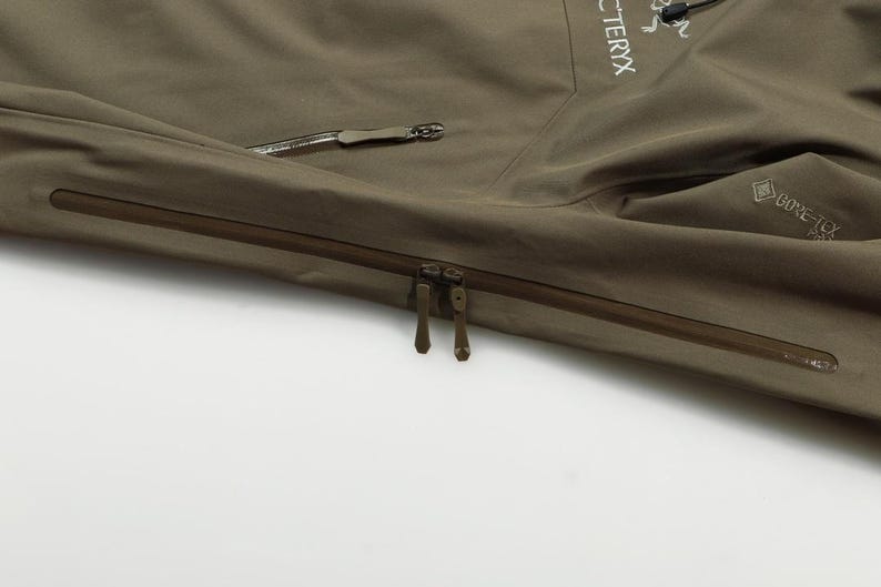 May include: Close-up of a khaki-colored jacket with a zipper closure and a side pocket. The jacket features the brand name "Arc'teryx" and the "Gore-Tex" logo. The jacket is made of a durable, waterproof material.