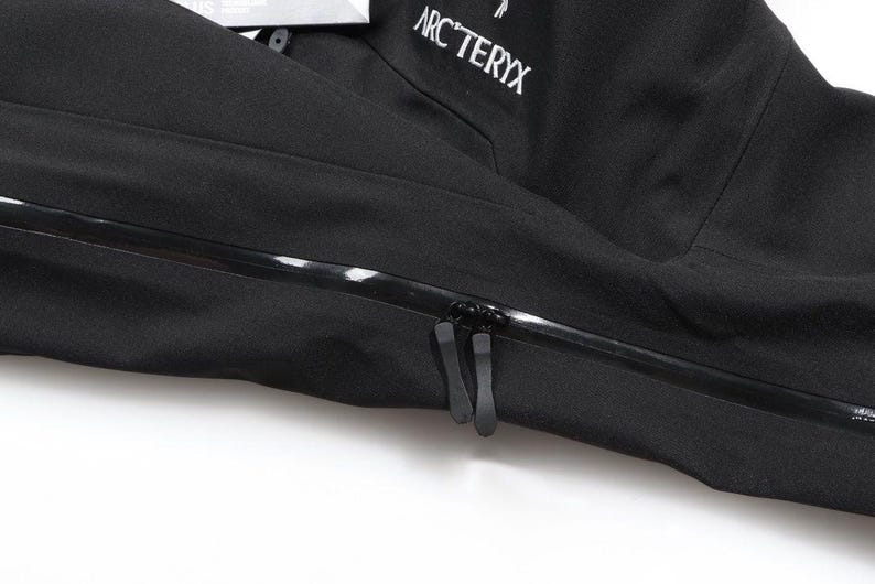 May include: Close-up of a black jacket with a prominent zipper and the brand name "ARC'TERYX" in white lettering. The jacket appears to be made of a durable, weather-resistant material, suitable for outdoor activities.