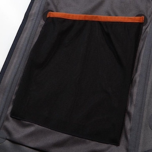 May include: Close-up of the interior of a dark gray jacket. A large black mesh pocket is visible, with an orange trim at the top. The jacket has a dark blue zipper and a gray lining.