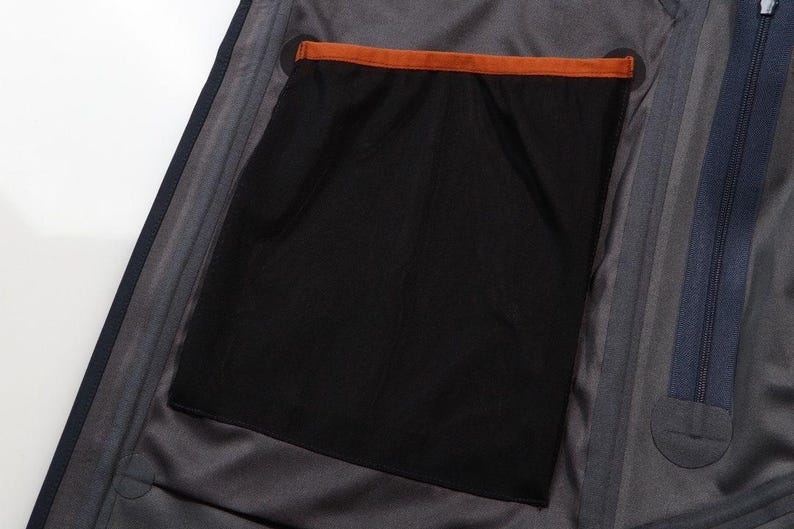 May include: Close-up of the interior of a dark gray jacket. A large black mesh pocket is visible, with an orange trim at the top. The jacket has a dark blue zipper and a gray lining.