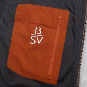 May include: Close-up of a jacket's interior, featuring a burnt orange pocket with a zipper and the white letters "B" over "SV". The pocket is set against a dark gray fabric, with a visible zipper and a portion of the jacket's lining.