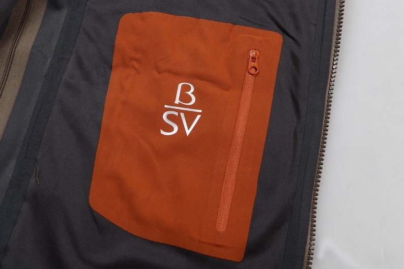 May include: Close-up of a jacket's interior, featuring a burnt orange pocket with a zipper and the white letters "B" over "SV". The pocket is set against a dark gray fabric, with a visible zipper and a portion of the jacket's lining.
