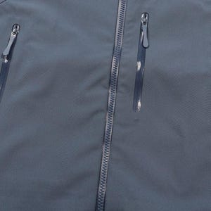 May include: Close-up of a slate blue jacket with three vertical zippered pockets and a central zipper. The zippers have silver pulls. The fabric has a subtle texture.