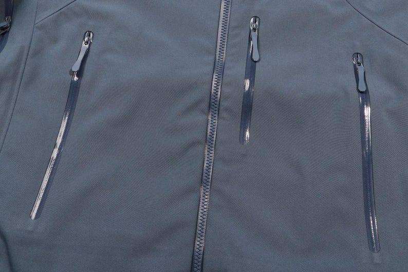 May include: Close-up of a slate blue jacket with three vertical zippered pockets and a central zipper. The zippers have silver pulls. The fabric has a subtle texture.