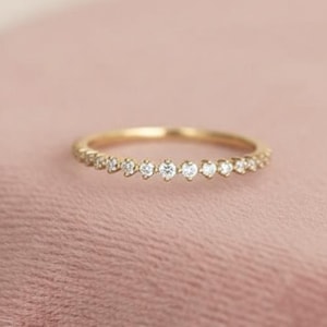 May include: A gold ring featuring a band of small diamonds. The ring is set against a soft pink backdrop, which accentuates the ring's design. The ring is a piece of jewellery. The ring's design is delicate.