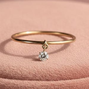 Könnte beinhalten: Ein zarter goldener Ring mit einem kleinen, hängenden, runden, klaren Edelstein. Der Ring ist vor einem weichen, rosa Hintergrund platziert. Das Band des Rings ist dünn und betont den Glanz des Edelsteins.