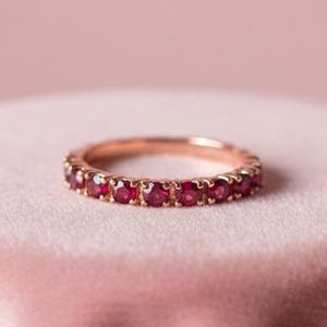 May include: A rose gold ring with a band of small, vibrant red gemstones. The ring is displayed on a pink velvet surface, set against a soft pink backdrop. The ring's design is simple and elegant, showcasing the rich color of the gemstones.