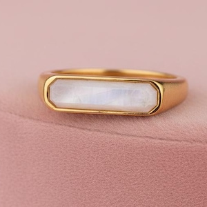 Puede incluir: Un anillo dorado con una piedra blanca rectangular e iridiscente. El anillo se muestra sobre una superficie rosa, creando una estética suave y elegante. El diseño del anillo es simple pero sofisticado, ideal para cualquier ocasión.