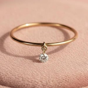 Pode incluir: Um delicado anel de ouro com um pequeno pendente de diamante. O anel é apresentado sobre um fundo rosa suave, realçando o seu design elegante. A fina banda do anel realça o brilho do diamante.