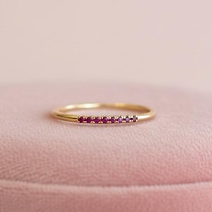 May include: A delicate gold ring featuring a row of small, vibrant red gemstones. The ring is set against a soft pink background, highlighting its elegant design. This piece of jewelry is a minimalist statement.