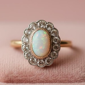 Puede incluir: Un anillo de oro con una piedra preciosa de ópalo ovalada, rodeada por un halo de pequeñas piedras preciosas transparentes. El anillo tiene un diseño vintage, con una banda de oro y un borde decorativo festoneado alrededor del halo.