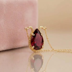 May include: A gold necklace featuring a pear-shaped, deep red gemstone pendant. The setting has a textured, ornate border with two small, curved gold elements extending from the top. The pendant hangs from a delicate, thin gold chain.