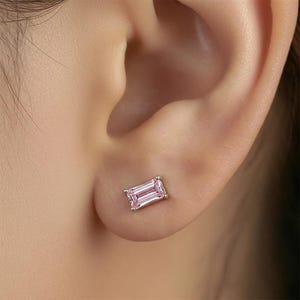 May include: A single stud earring featuring a rectangular, emerald-cut light pink gemstone set in a polished silver-toned metal prong setting, positioned securely on an earlobe. The stone displays clear facets that reflect light across its surface.