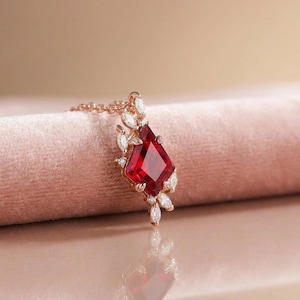 May include: A rose gold necklace featuring a kite-shaped red gemstone pendant. The central red stone is surrounded by small, marquise-cut clear accent stones arranged in a cluster along the edges of the setting, attached to a delicate chain.