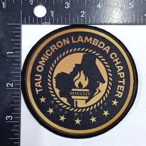 May include: A round patch with a black background and gold trim. The patch features the text "TAU OMICRON LAMBDA CHAPTER" in gold around the edge. Inside is a gold map, a torch, and the Roman numeral "MMXXIV".