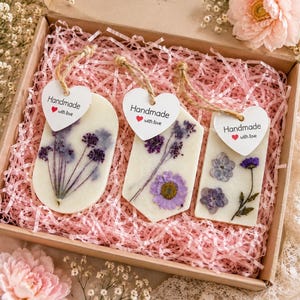 May include: Three white wax tablets with embedded dried purple flowers, each featuring a heart-shaped tag reading Handmade with love. The tablets are oval, pentagonal, and rectangular, resting on pink shredded paper inside a cardboard gift box.