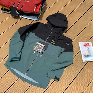 May include: A teal and black hooded jacket with a zipper closure and the brand name "Arc'teryx" visible. The jacket has a "GORE-TEX" tag attached. Several other jackets are stacked in the background.