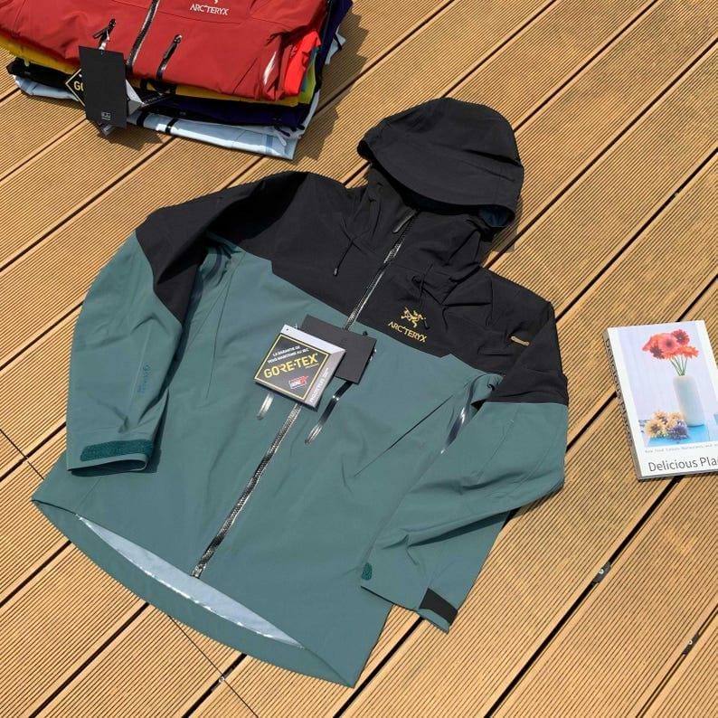 May include: A teal and black hooded jacket with a zipper closure and the brand name "Arc'teryx" visible. The jacket has a "GORE-TEX" tag attached. Several other jackets are stacked in the background.