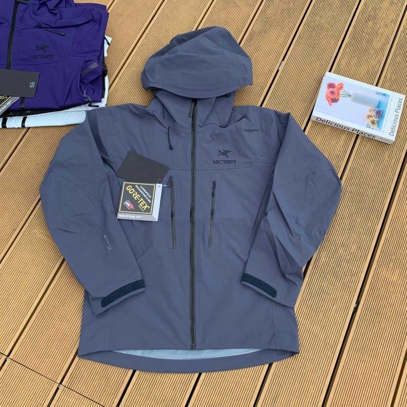 May include: A gray hooded jacket with a full-length zipper, two front pockets, and the "Arc'teryx" logo. A "GORE-TEX" tag is attached to the jacket. Another purple jacket is visible in the background.