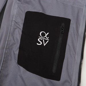 May include: Close-up of a gray jacket interior with a black pocket. The pocket features a white logo and a black zipper. The jacket has a full-length black zipper and a gray lining.