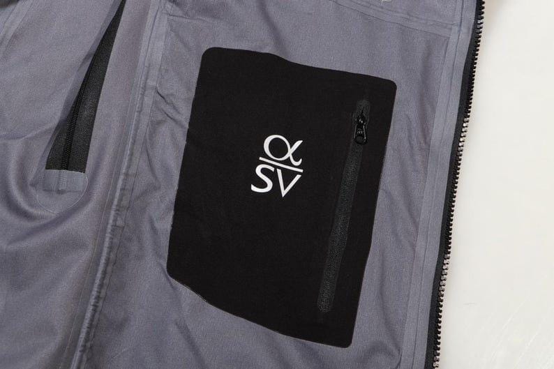 May include: Close-up of a gray jacket interior with a black pocket. The pocket features a white logo and a black zipper. The jacket has a full-length black zipper and a gray lining.