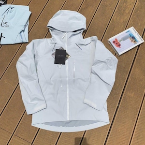 May include: A light blue hooded jacket with a full-length zipper and two front pockets. The jacket has a tag attached and is laid out on a wooden surface. A light blue shirt is visible in the background.