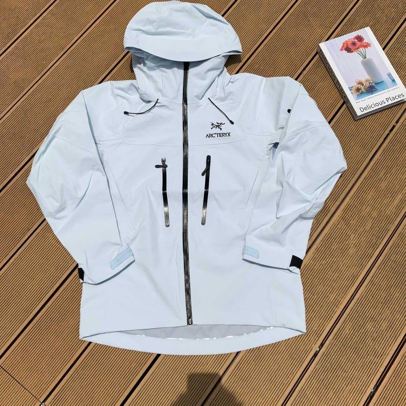 May include: A light blue hooded jacket with a full-length zipper and multiple pockets. The jacket features the brand name "Arc'teryx" on the chest. The jacket is on a wooden surface.