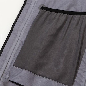 May include: Close-up of a gray garment interior with a large, dark gray mesh pocket. The pocket is outlined with black trim and is sewn into the gray fabric. The garment has a black trim along the edge.