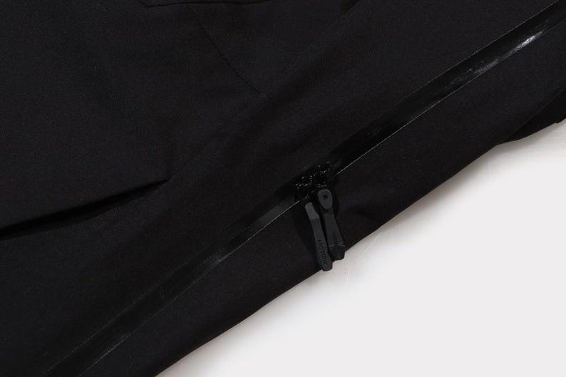 May include: Close-up of a black garment with a visible zipper. The zipper is black and has two pulls. The fabric appears to be a smooth, matte material. The image is set against a white background.