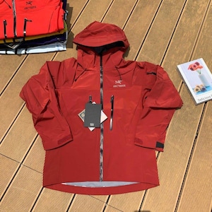 May include: A red hooded jacket with a full-length zipper, two side pockets, and the brand name "Arc'teryx" on the chest. The jacket has a tag attached and is displayed on a wooden surface. Additional jackets are stacked in the background.