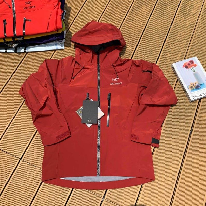 May include: A red hooded jacket with a full-length zipper, two side pockets, and the brand name "Arc'teryx" on the chest. The jacket has a tag attached and is displayed on a wooden surface. Additional jackets are stacked in the background.