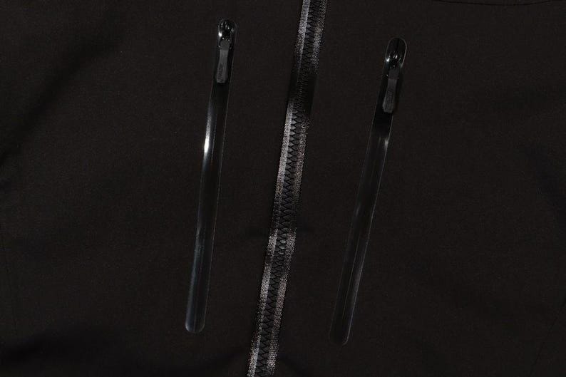 May include: Close-up of a black garment featuring three zippers. Two long, thin zippers flank a central zipper with a silver-colored track. The zippers are all black with silver-colored teeth.