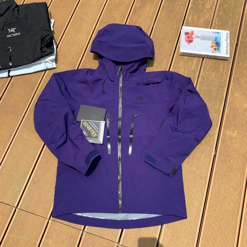 May include: A vibrant purple Arc'teryx Gore-Tex jacket with a hood, zipper closure, and multiple pockets. The jacket has the Arc'teryx logo on the chest and sleeve. A tag with the Gore-Tex logo is attached. Additional black jacket is visible.