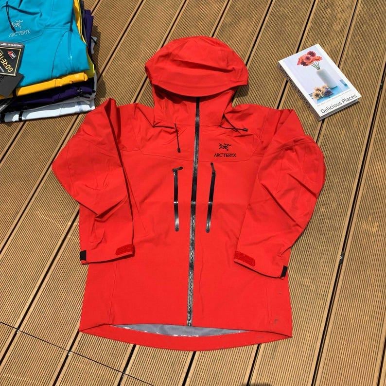 May include: A bright red hooded jacket with a full-length zipper and the brand name "Arc'teryx" on the chest. The jacket has multiple pockets and adjustable cuffs. A book titled "Delicious Places" is on the right side of the jacket.