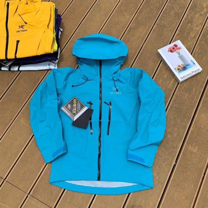 May include: A turquoise hooded jacket with a black zipper and two front pockets. The jacket has the brand name "Arc'teryx" on the chest and a "GORE-TEX" label. Additional jackets in yellow and purple are stacked in the background.