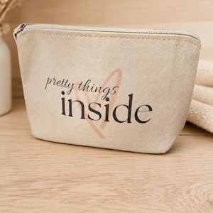 Könnte beinhalten: Ein beigefarbenes Reißverschlusstäschchen mit dem in Schwarz aufgedruckten Text "pretty things inside" und einem blassen, herzförmigen Umriss hinter dem Schriftzug. Das Täschchen hat eine einfache, rechteckige Form mit abgerundeten Ecken.
