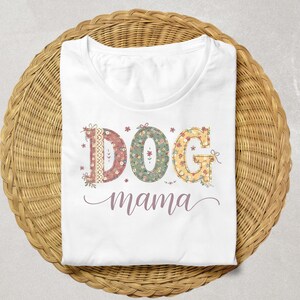Op de afbeelding: Wit T-shirt met de woorden "DOG mama" in een bloemenontwerp. Het woord "DOG" staat in afzonderlijke letters met verschillende bloempatronen en kant. Het woord "mama" staat in een cursief lettertype. Het shirt is opgevouwen op een geweven, cirkelvormig oppervlak.