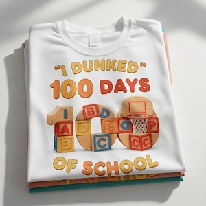 Puede incluir: Camiseta blanca con el texto "I Dunked 100 Days of School" en naranja y marrón. El diseño incluye una canasta de baloncesto y bloques de madera con letras y números. La camiseta está doblada encima de otras camisetas.