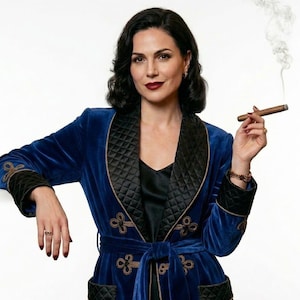 May include: A woman wearing a blue velvet robe with black quilted lapels and gold embroidery. The robe is belted at the waist. She is holding a lit cigar.