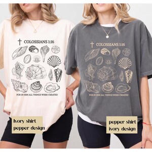 Könnte beinhalten: Zwei kurzärmelige T-Shirts mit Rundhalsausschnitt, eines in Elfenbein mit schwarzem Aufdruck und eines in Pfeffergrau mit elfenbeinfarbenem Aufdruck. Beide zeigen ein Kreuz, den Text COLOSSIANS 1:16, Muschelillustrationen und den Text FOR IN HIM ALL THINGS WERE CREATED.
