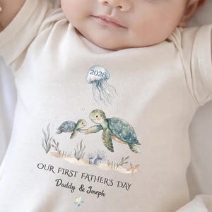 May include: Cream-colored baby onesie featuring a watercolor design of two sea turtles and a jellyfish. The text "OUR FIRST FATHER'S DAY Daddy & Joseph" is printed below the turtles. The jellyfish has the number "2026" on it.