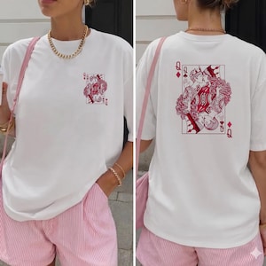 May include: White t-shirt featuring a red and white Queen of Diamonds playing card design on the front and back. The shirt is worn with pink and white striped shorts. A gold chain necklace and bracelet are visible.