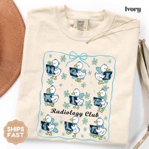 Könnte beinhalten: Elfenbeinfarbenes T-Shirt mit einer Grafik von Cartoon-Enten, die Röntgenbilder halten, umgeben von einem blauen Blumenrahmen. Der Text "Radiology Club" steht darunter. Das Shirt hat ein Comfort Colors-Etikett.
