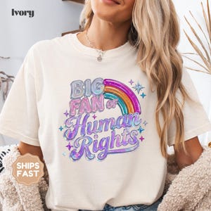 May include: Ivory-colored t-shirt with the text "BIG FAN of Human Rights" in a colorful, holographic design. The design includes a rainbow and star accents. The shirt is a casual, short-sleeved style.