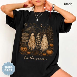 Pode incluir: Uma t-shirt preta com uma estampa gráfica de três fantasmas num campo com um celeiro, abóboras e uma placa com os dizeres Pumpkins, Hay Rides e Corn Maze. O texto 'tis the season' está impresso em letra cursiva na parte inferior.