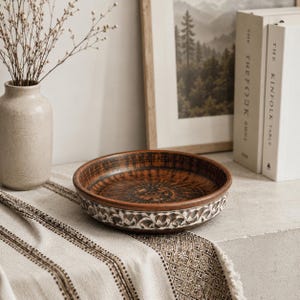 Könnte beinhalten: Eine runde, braune Keramikschale mit dekorativem weißem Rand. Die Schale hat ein detailliertes, gemustertes Innendesign und liegt auf einem gemusterten Textil. Eine Vase mit Trockenblumen und Bücher befinden sich im Hintergrund.