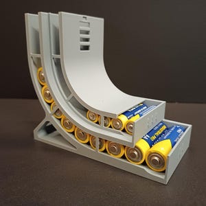 Op de afbeelding: Een grijze 3D-geprinte batterijorganizer met meerdere AA-batterijen. De organizer heeft een gebogen ontwerp met meerdere sleuven om de batterijen in op te bergen. De batterijen zijn geel en zilver met blauwe labels.