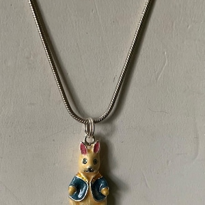 May include: A silver chain necklace with a pendant featuring a small, yellow rabbit wearing a blue jacket. The rabbit has pink ears and a black nose and mouth. The pendant is approximately 1 inch in height.