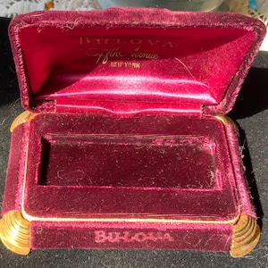 May include: A red velvet jewelry box with gold trim and the words "Bulova Fifth Avenue New York" on the inside of the lid. The box is open and the inside is lined with red velvet.